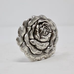 Puede incluir: Objeto decorativo plateado con forma de rosa. Los p&eacute;talos est&aacute;n intrincadamente tallados, creando un dise&ntilde;o floral realista. Probablemente sea un elemento decorativo para la decoraci&oacute;n del hogar o joyer&iacute;a.