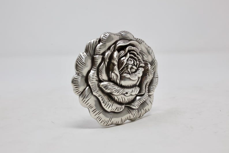 Puede incluir: Objeto decorativo plateado con forma de rosa. Los p&eacute;talos est&aacute;n intrincadamente tallados, creando un dise&ntilde;o floral realista. Probablemente sea un elemento decorativo para la decoraci&oacute;n del hogar o joyer&iacute;a.