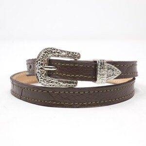 May include: A brown leather belt with a decorative silver buckle and a matching silver tip. The belt is stitched with a lighter brown thread and is wrapped around itself.
