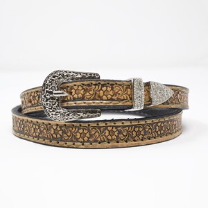 May include: A brown leather belt with a floral embossed design. The belt has a silver-colored filigree buckle and a smaller, decorative buckle with rhinestones. The belt is likely for fashion or Western wear.