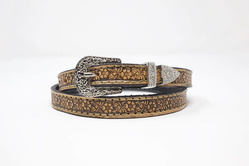 May include: A brown leather belt with a floral embossed design. The belt has a silver-colored filigree buckle and a smaller, decorative buckle with rhinestones. The belt is likely for fashion or Western wear.