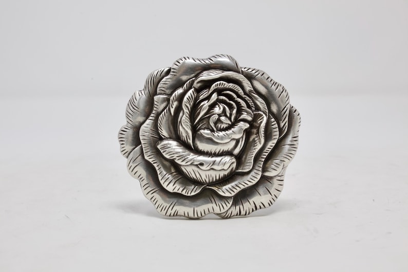 Puede incluir: Escultura de rosa plateada con p&eacute;talos intrincadamente tallados. El dise&ntilde;o floral realista se destaca sobre un fondo blanco liso, resaltando la forma y la textura. Esta pieza decorativa se puede usar como broche o elemento decorativo.