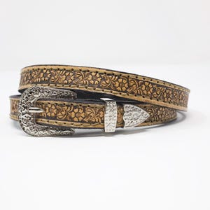 May include: A tan leather belt with a floral embossed pattern. The belt has a silver-colored buckle and a matching belt loop. The belt is rolled up on a white background.