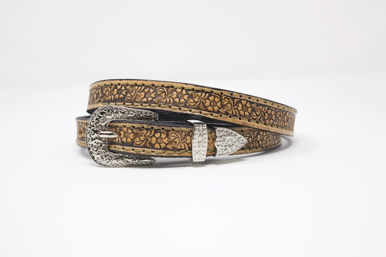 May include: A tan leather belt with a floral embossed pattern. The belt has a silver-colored buckle and a matching belt loop. The belt is rolled up on a white background.