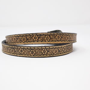 May include: A brown leather belt with a floral pattern. The belt is coiled, showing the intricate carved design. The edges are dark, and the floral pattern is a lighter brown, creating contrast. The belt has a buckle.
