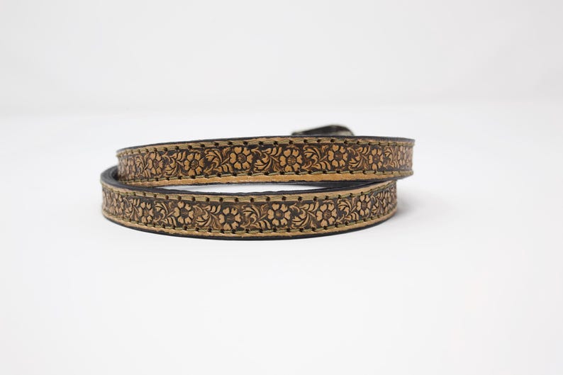May include: A brown leather belt with a floral pattern. The belt is coiled, showing the intricate carved design. The edges are dark, and the floral pattern is a lighter brown, creating contrast. The belt has a buckle.