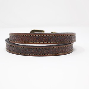 May include: A brown leather bracelet with a floral pattern. The bracelet is wrapped around itself, showing the intricate tooling and stitching. The buckle is a gold color. The bracelet is a stylish accessory.