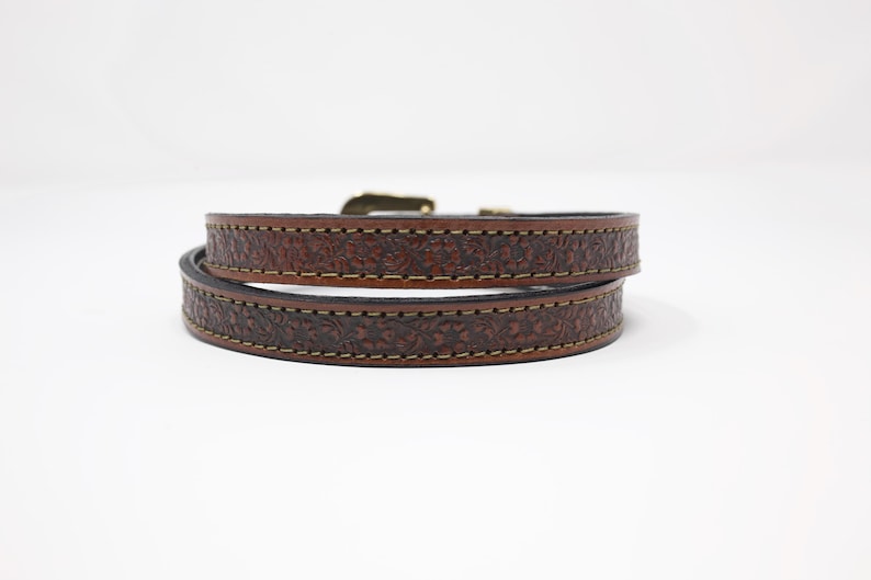 May include: A brown leather bracelet with a floral pattern. The bracelet is wrapped around itself, showing the intricate tooling and stitching. The buckle is a gold color. The bracelet is a stylish accessory.