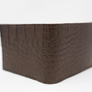 May include: A dark brown bifold wallet featuring a textured crocodile skin pattern on the exterior surface. The wallet is constructed with visible stitching along the top edge, showcasing a rectangular shape and a deep, rich brown color throughout.