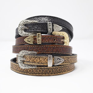 May include: A stack of leather belts with ornate silver and gold buckles. The belts are in various shades of brown and black, with embossed floral patterns. The buckles have detailed designs.