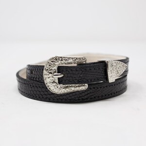 May include: Black belt with a textured pattern and silver-colored buckle and tip. The buckle has a decorative design. The belt is rolled up, showing the buckle and tip. The belt is on a white background.