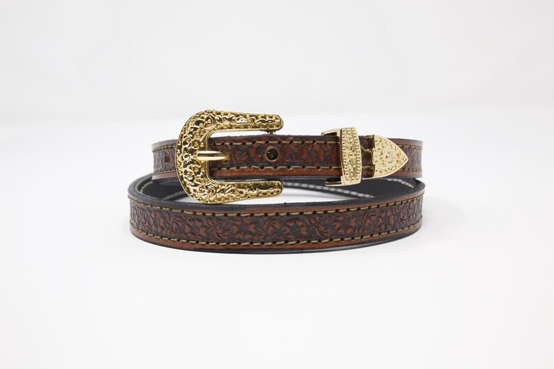 May include: A brown leather belt with a gold-colored buckle and floral embossed design. The belt has a gold-colored buckle and a matching gold-colored belt tip. The belt is approximately 1 inch wide.