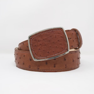 May include: A brown leather belt with a textured surface and a large, rectangular buckle with a silver-colored frame. The belt is rolled up, showcasing its full length and design.
