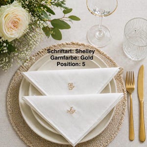 Könnte beinhalten: Zwei weiße Stoffservietten, die zu Dreiecken gefaltet sind und jeweils ein kleines, goldfarben besticktes Monogramm aufweisen. Die Servietten liegen auf einem weißen Keramikteller. Der Text lautet: Schriftart: Shelley, Garnfarbe: Gold, Position: 5.