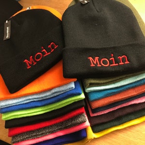Könnte beinhalten: Ein Stapel bunter Strickmützen mit dem Wort "Moin" auf der Vorderseite gestickt. Die Mützen sind schwarz, orange, blau, grün, rot und gelb.