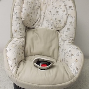 Puede incluir: Un asiento de coche para bebé beige con una funda de tela de estampado de llama blanco y marrón. El asiento tiene un arnés de cinco puntos y un reposacabezas incorporado.