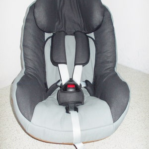 Puede incluir: Un asiento de coche gris y negro con un arnés de cinco puntos. El asiento tiene un reposacabezas acolchado y una cubierta de tela gris suave.