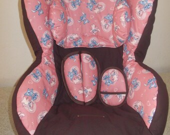 May include: A child's car seat with a brown exterior and pink interior. The pink fabric is patterned with blue cartoon characters. The seat has a harness and headrest for safety. The seat is designed to be installed in a vehicle.