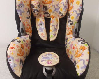 May include: A black car seat cover with a cream background and a pattern of cartoon cats. The cover has a headrest, shoulder pads, and a crotch strap.