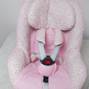 Puede incluir: Silla de coche rosa para bebé con estampado floral blanco y arnés de cinco puntos gris.