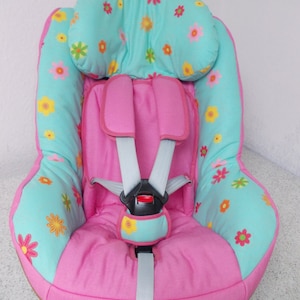 Puede incluir: Un asiento de coche rosa y turquesa con un patrón floral. El asiento tiene un arnés de cinco puntos y un reposacabezas.