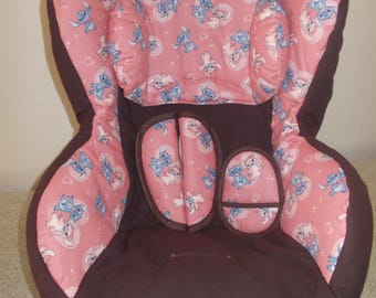 May include: A child's car seat with a brown exterior and pink interior. The pink fabric is patterned with blue cartoon characters. The seat has a harness and headrest for safety. The seat is designed to be installed in a vehicle.