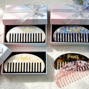 May include: Five semi-circular hair combs made of patterned acrylic resin, each personalized with a name in gold or white script. Three combs are displayed inside light blue and pink gift boxes with silver ribbons, while two rest outside.