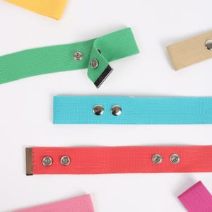 May include: A collection of colorful canvas belts in shades of green, blue, red, pink, yellow, and tan. Each belt features metal hardware and snap closures. The belts are arranged on a white background.