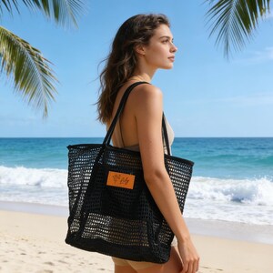 K&ouml;nnte beinhalten: Eine schwarze Netztasche mit einem rechteckigen orangefarbenen Etikett. Die Tasche wird &uuml;ber der Schulter an einem Strand mit Palmen und dem Ozean im Hintergrund getragen. Geeignet f&uuml;r Strandutensilien.