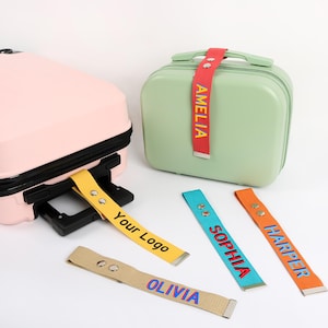 May include: A light pink suitcase and a light green suitcase with colorful luggage tags. The tags are yellow, red, blue, orange, and tan, with names and the text "Your Logo" printed on them. The tags are attached to the suitcases.