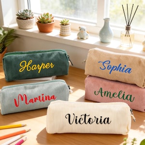 Op de afbeelding: Vijf corduroy etuis in verschillende kleuren: groen, beige, lichtblauw, roze en crème. Elke etui heeft een geborduurde naam: Harper, Sophia, Marina, Amelia en Victoria. De etuis hebben een ritssluiting en liggen op een houten oppervlak.