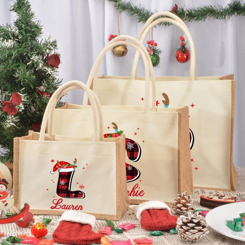 May include: Three beige burlap tote bags with red and black plaid Christmas stocking designs. The smallest bag has the name "Lauren" printed on it, the middle bag has the name "Jackie" printed on it, and the largest bag has a reindeer design.