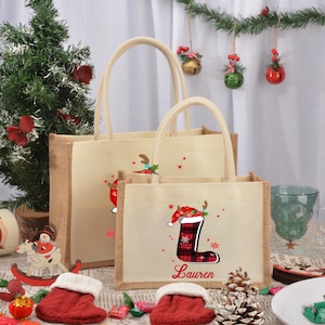 May include: Two burlap tote bags with Christmas designs. The smaller bag has a red and black plaid stocking with a Santa hat on top and the name "Lauren" printed below. The larger bag has a similar design with a reindeer.