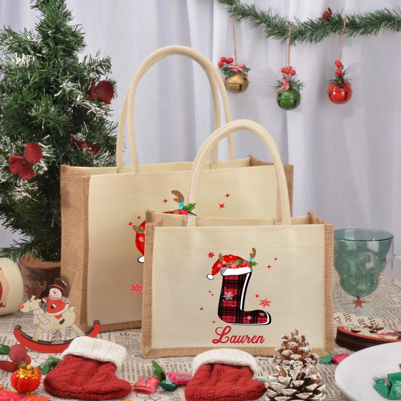 May include: Two burlap tote bags with Christmas designs. The smaller bag has a red and black plaid stocking with a Santa hat on top and the name "Lauren" printed below. The larger bag has a similar design with a reindeer.