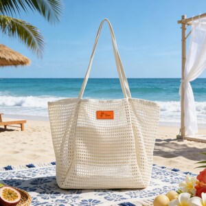 K&ouml;nnte beinhalten: Eine gro&szlig;e, cremefarbene Netztasche mit langen Henkeln, die auf einer gemusterten Stranddecke liegt. Die Tasche hat ein kleines orangefarbenes Etikett. Der Hintergrund zeigt eine Strandszene mit einem Strohschirm und dem Ozean.