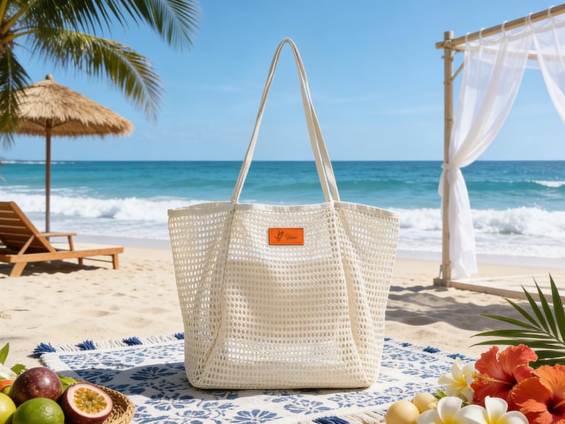 K&ouml;nnte beinhalten: Eine gro&szlig;e, cremefarbene Netztasche mit langen Henkeln, die auf einer gemusterten Stranddecke liegt. Die Tasche hat ein kleines orangefarbenes Etikett. Der Hintergrund zeigt eine Strandszene mit einem Strohschirm und dem Ozean.