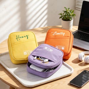 May include: Three square zippered travel pouches in yellow, orange, and purple. The yellow pouch features the name Henry, the orange pouch features the name Emily, and the purple pouch contains a white power bank and charging cables.