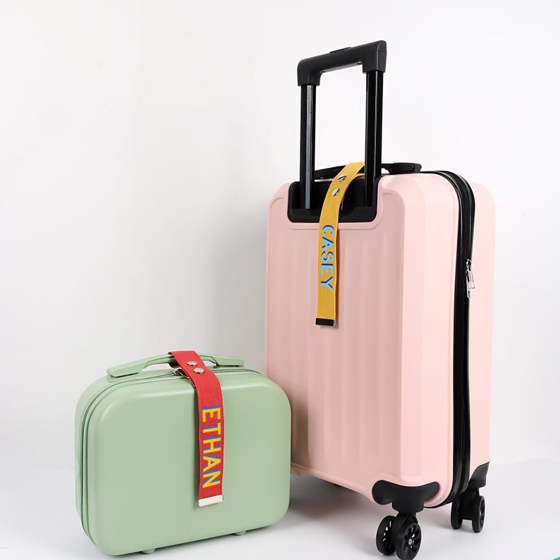 May include: A light pink rolling suitcase with a black handle and zipper, next to a small, light green travel case. Both have colorful luggage tags with names. The larger suitcase has a yellow tag that says "CASLY". The smaller case has a red tag that says "ETHAN".