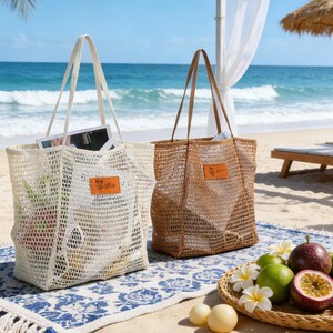K&ouml;nnte beinhalten: Zwei gewebte Tragetaschen mit Henkeln, eine cremefarbene und eine braune, liegen am Strand. Jede Tasche hat ein rechteckiges Etikett mit Text. Die Taschen sind offen und zeigen den Inhalt. Ein blau-wei&szlig; gemusterter Teppich und ein Obstteller befinden sich im Vordergrund.
