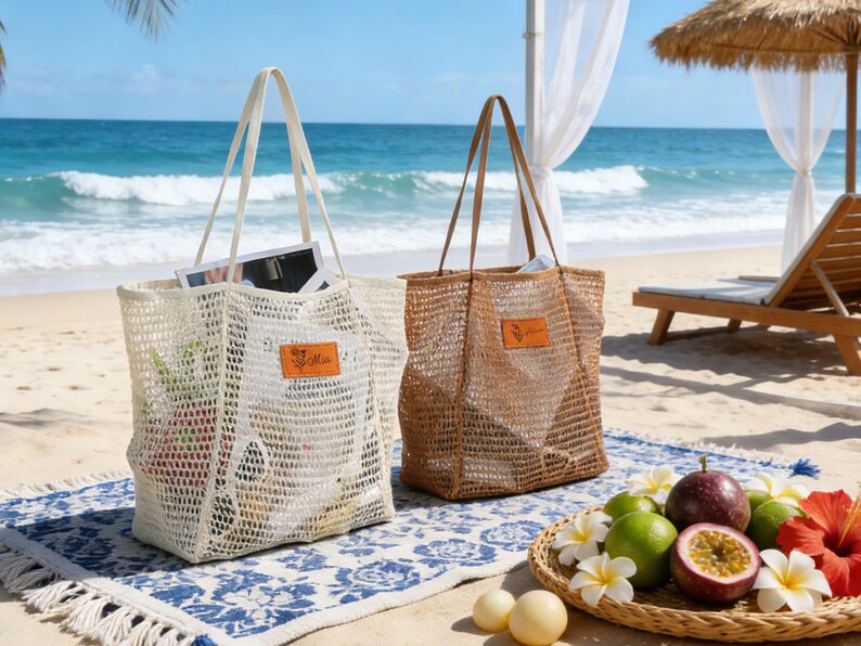 K&ouml;nnte beinhalten: Zwei gewebte Tragetaschen mit Henkeln, eine cremefarbene und eine braune, liegen am Strand. Jede Tasche hat ein rechteckiges Etikett mit Text. Die Taschen sind offen und zeigen den Inhalt. Ein blau-wei&szlig; gemusterter Teppich und ein Obstteller befinden sich im Vordergrund.
