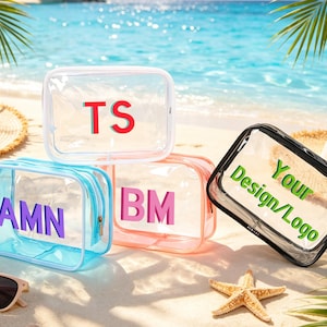 Op de afbeelding: Transparante cosmetische tassen met gepersonaliseerde initialen in rood, paars en roze. Eén tas heeft de tekst "Your Design/Logo" in groen. De tassen liggen op een zandstrand met zonnebrillen en hoeden.