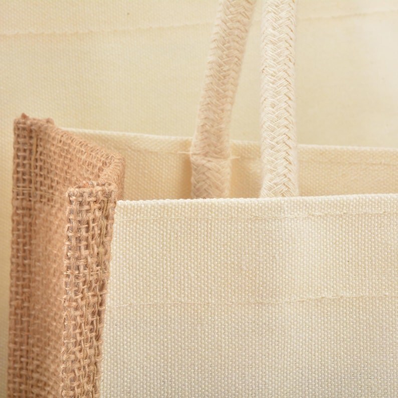 May include: A close-up of a natural-colored canvas tote bag with a natural-colored jute bag peeking out from behind it. The tote bag has two thick cotton rope handles.