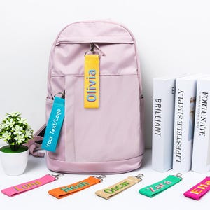Könnte beinhalten: Ein helllila Rucksack mit einem gelben Anhänger mit der Aufschrift "Olivia" und einem blauen Anhänger mit "Your Text/Logo". Mehrere bunte Schlüsselanhänger mit Namen werden vor dem Rucksack angezeigt.