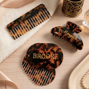 May include: A set of three hair accessories with a brown and black tortoiseshell pattern. One rectangular comb is engraved with the name Katie, one claw clip is engraved with the name Morgan, and one oval compact comb is engraved with the name BROOKE.