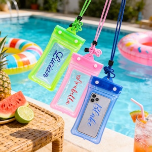 Puede incluir: Tres fundas impermeables para teléfono en verde, rosa y azul, cada una con un cordón. La funda verde tiene el nombre "Lucian", la rosa "Arabella" y la azul "Blake". El fondo muestra una piscina, fruta y una bebida.