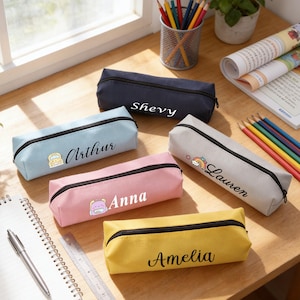 Puede incluir: Una colección de estuches personalizados en varios colores, incluyendo azul, rosa, amarillo y gris. Cada estuche presenta una cremallera negra y un nombre impreso en una fuente cursiva. Algunos estuches tienen pequeñas imágenes decorativas.