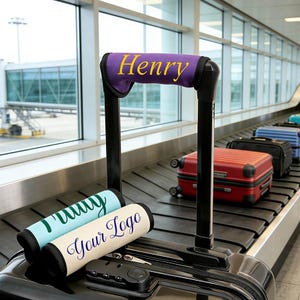 May include: Luggage handle wraps in various colors with custom text. One wrap is purple with "Henry" in yellow. Other wraps are light blue with "Tommy" and cream with "Your Logo". Luggage is on a baggage carousel.