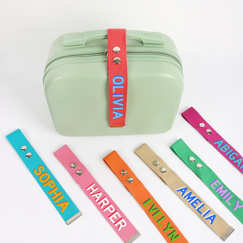 May include: A light green suitcase with a red strap labeled "OLIVIA". Several colorful luggage tags with names like "SOPHIA", "HARPER", and "AMELIA" are scattered around the suitcase. The tags are in various colors, including blue, pink, orange, and green.