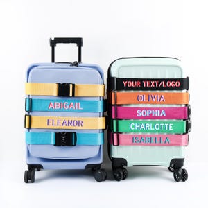 May include: Two rolling suitcases, one light blue and one mint green, each secured with colorful luggage straps. The straps display names like "Abigail," "Eleanor," "Olivia," and "Sophia." The straps have black buckles.