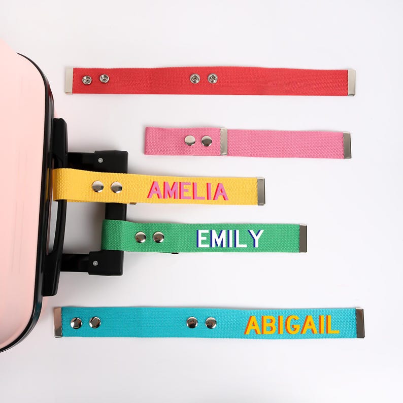 May include: A collection of colorful luggage straps with metal hardware. The straps are red, pink, yellow, green, and teal, each with a name printed in a coordinating color: Amelia, Emily, and Abigail. The straps are attached to a pink suitcase.